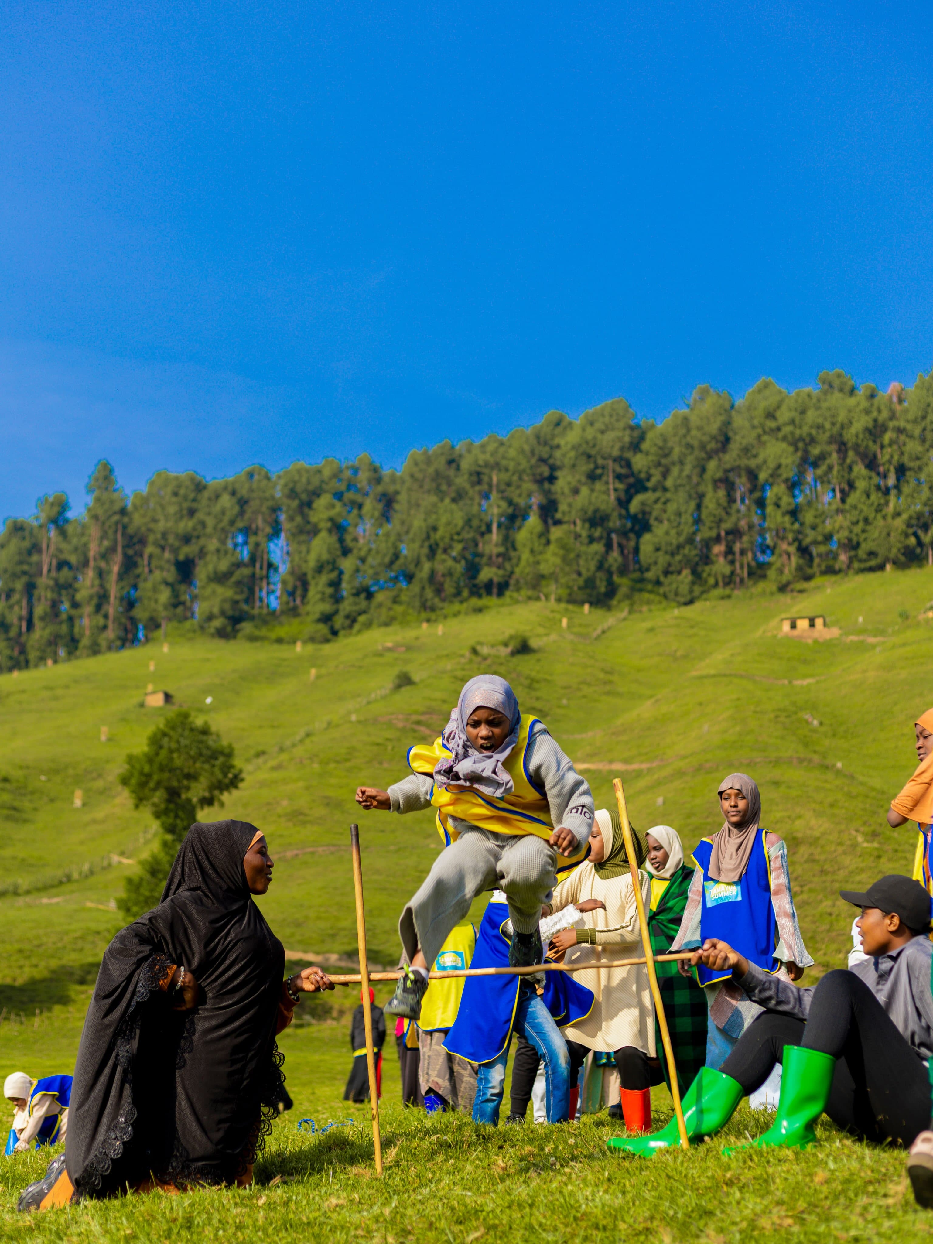 A Tamayuz Summer Camp student jumping over a stick game, cheered on by peers